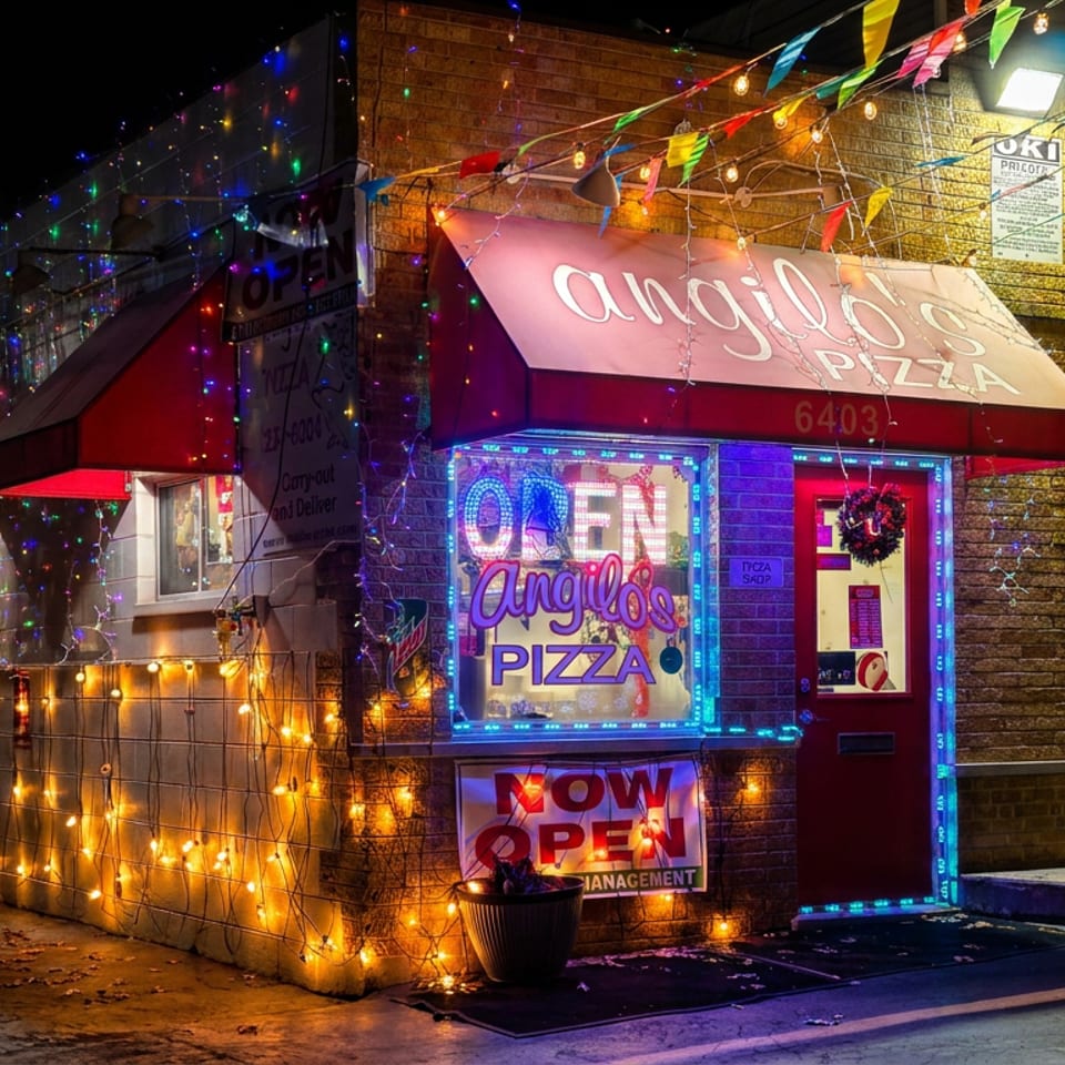 Angelo’s Pizza storefront glowing with colorful string lights, festive pennants, red awnings, and illuminated signage at night.